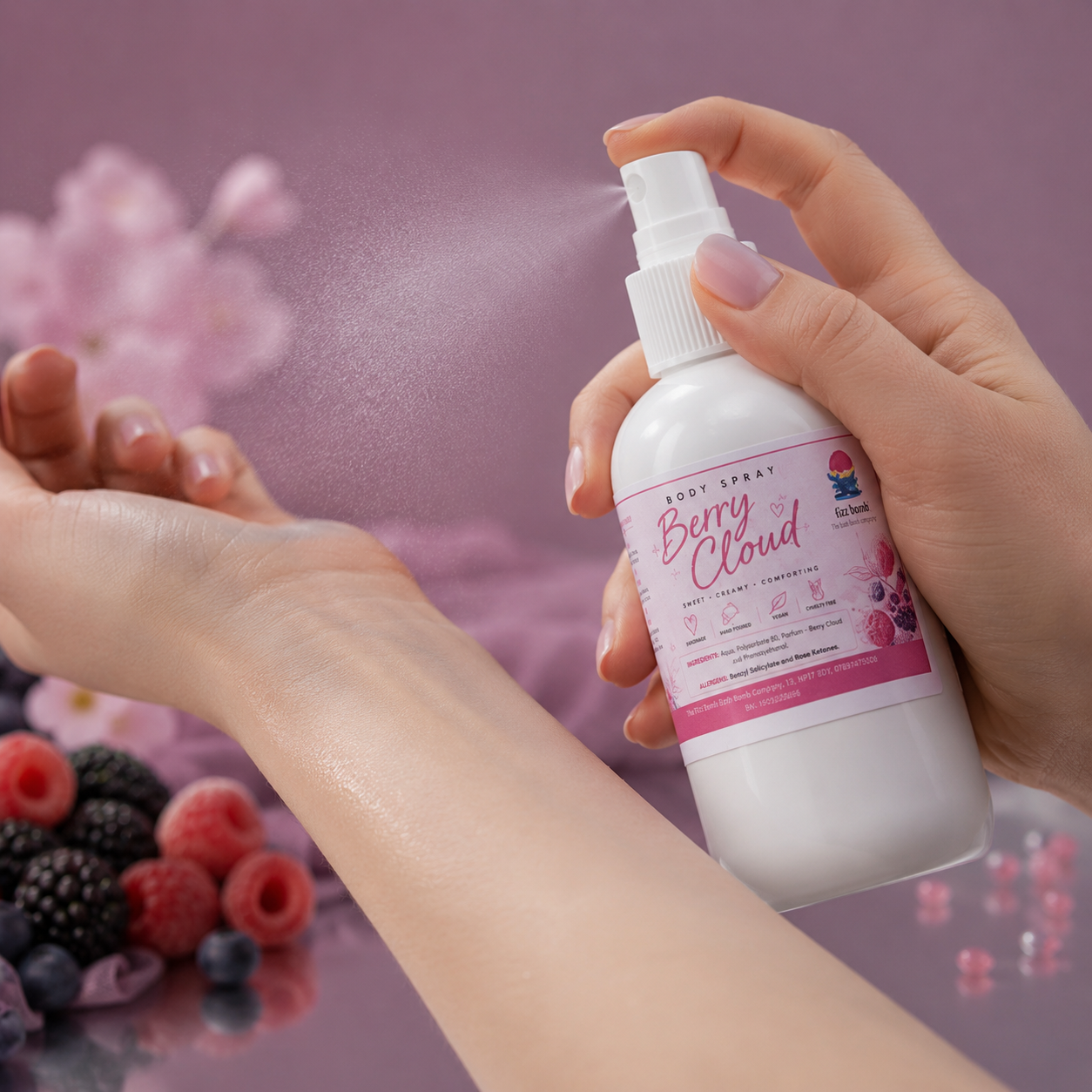 A hand holds a white body spray bottle labeled Berry Cloud while spraying a fine mist onto the back of another hand. The background is a soft pink with blurred berries and floral elements.