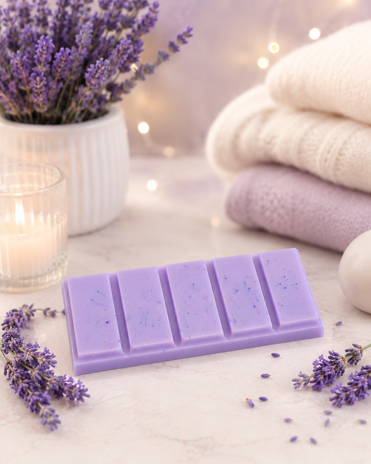 A lavender-colored segmented wax bar with tiny blue flecks, styled on a marble surface with lavender sprigs, a candle, a white pot of lavender, and folded towels in the background.